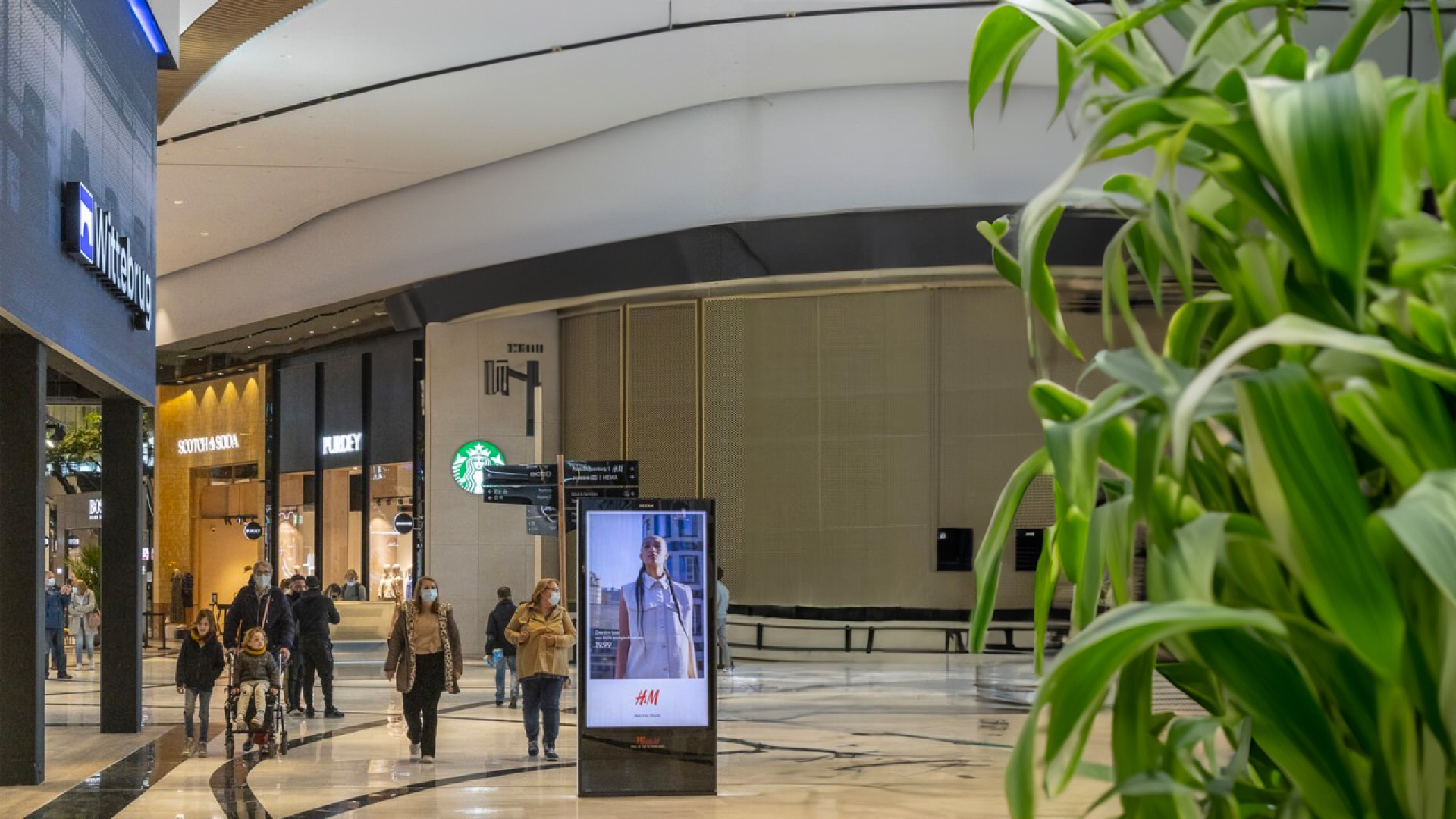 A shopping mall with people walking and a digital ad display in the center.