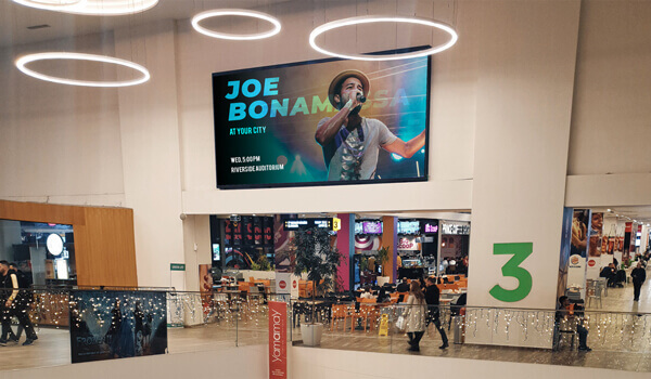 A DOOH screen installed inside a shopping mall promotes a concert event with the image of a celebrity and a call to action
