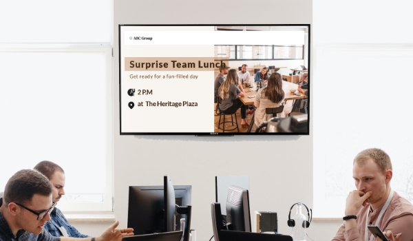 A digital screen installed in a workplace shows the message 'Surprise Team Lunch'